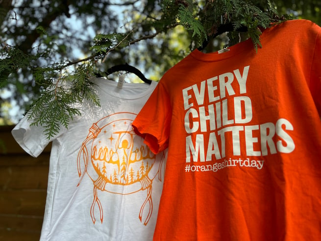 A shirt with an orange design featuring a circle, sacred pipe and trees by artist Miigizii hangs on cedar boughs next to an orange shirt with the words "Every Child Matters" printed in white