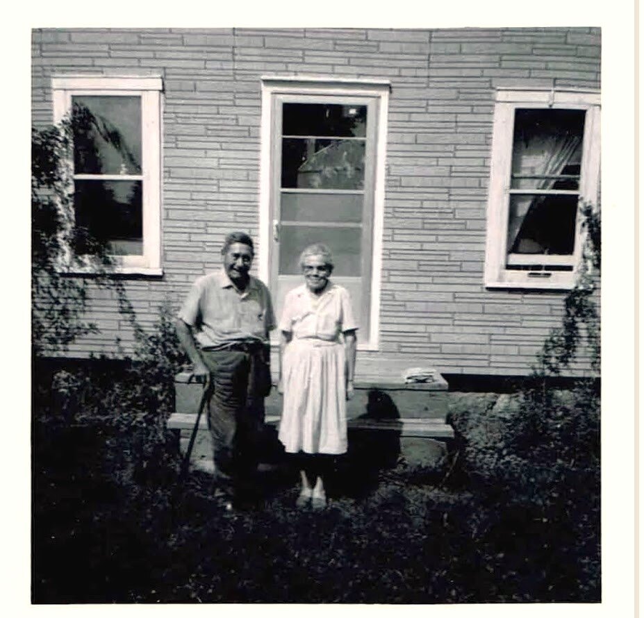 A black and white photo of an elderly couple, William holding a cane, and Lena Bomberry, wearing a white dress with short sleeves taken in 1968 in front of their home