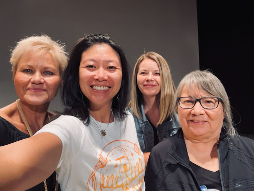 four women stand smiling together. Amanda has shoulder length blond hair.  she wears a black shirt and jean jacket.